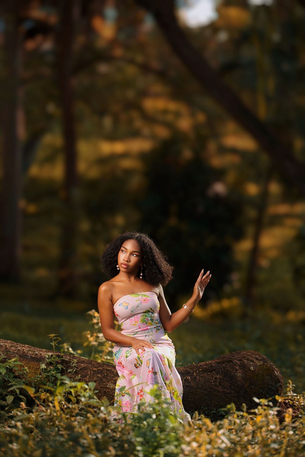 A woman in a floral dress poses gracefully on a fallen log in a sunlit forest setting during golden hour.