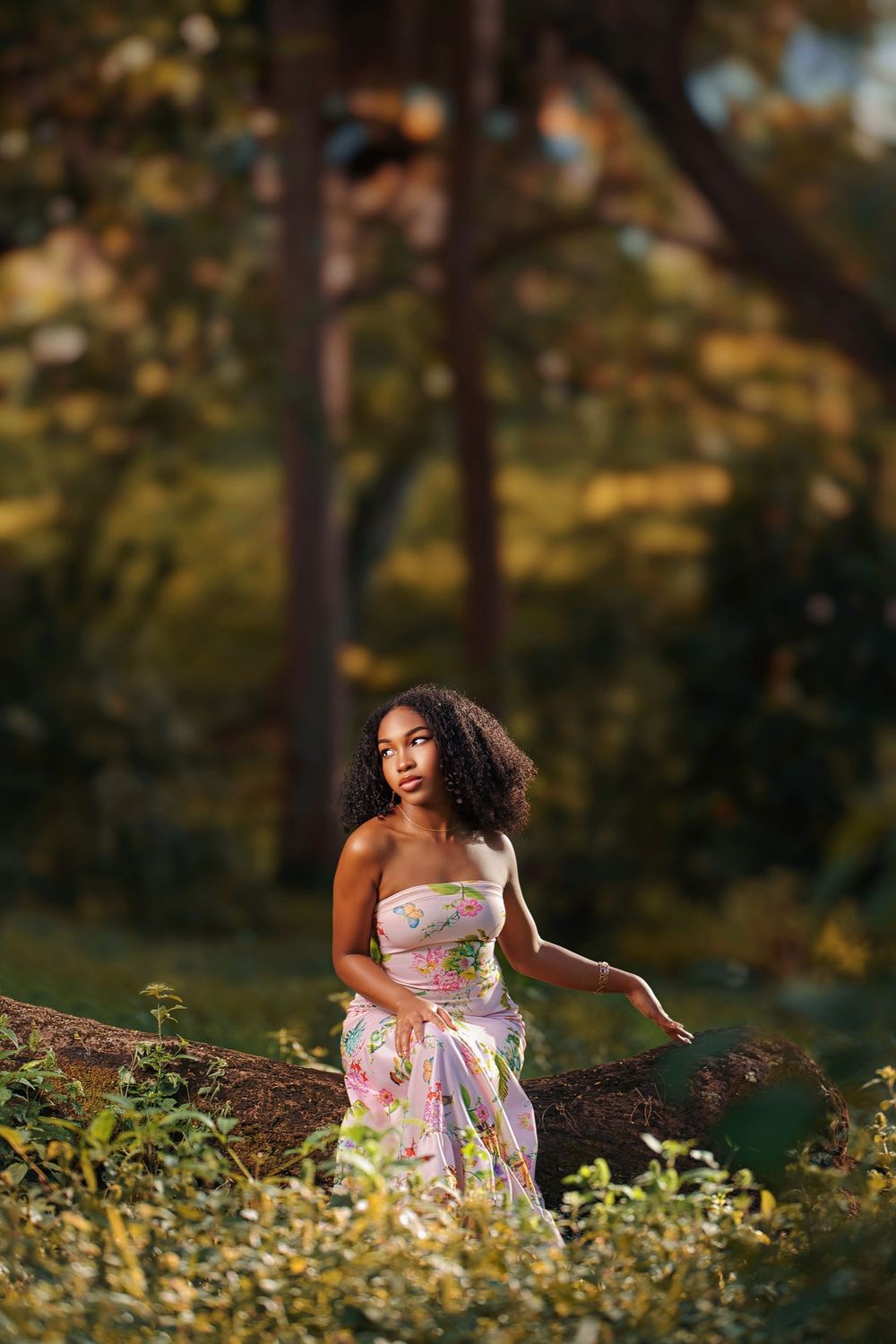 A person in a floral dress poses gracefully in a sunlit garden setting during golden hour.