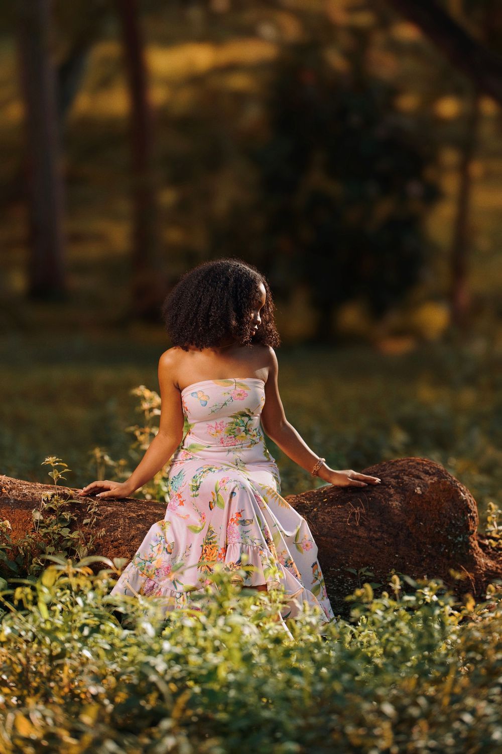 A person in a floral strapless dress sits on a log during golden hour in a garden setting.