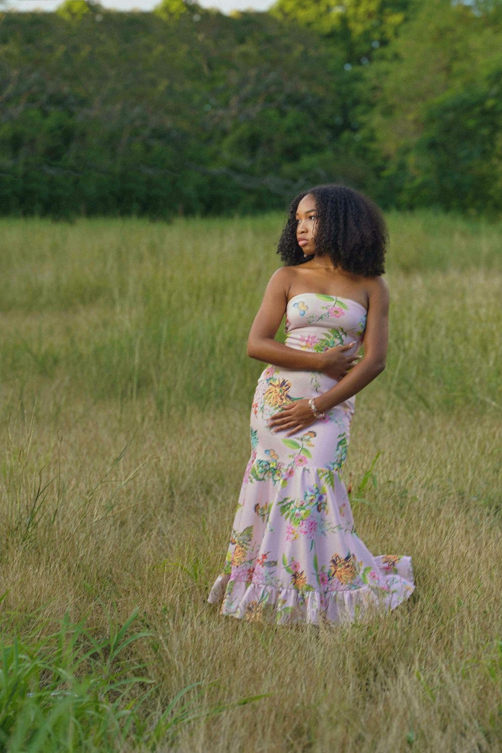 A person in a floral sundress poses gracefully in a sunlit meadow with tall grass and trees in the background.