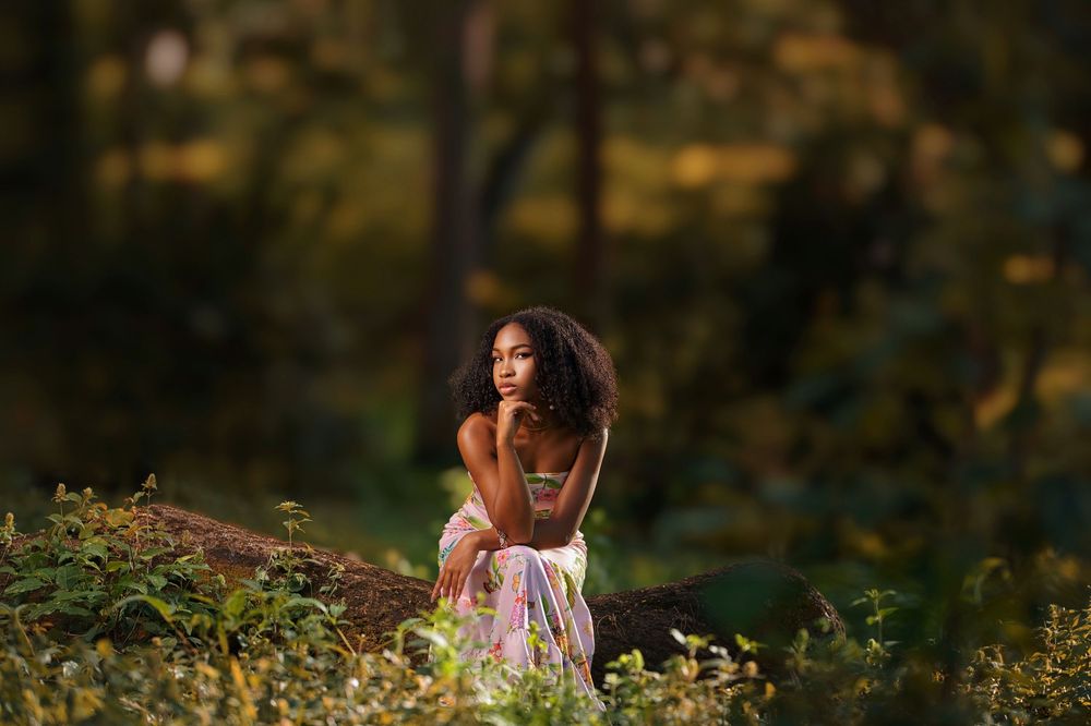 A person in a flowing dress poses thoughtfully in a sunlit meadow surrounded by wildflowers and trees at golden hour.
