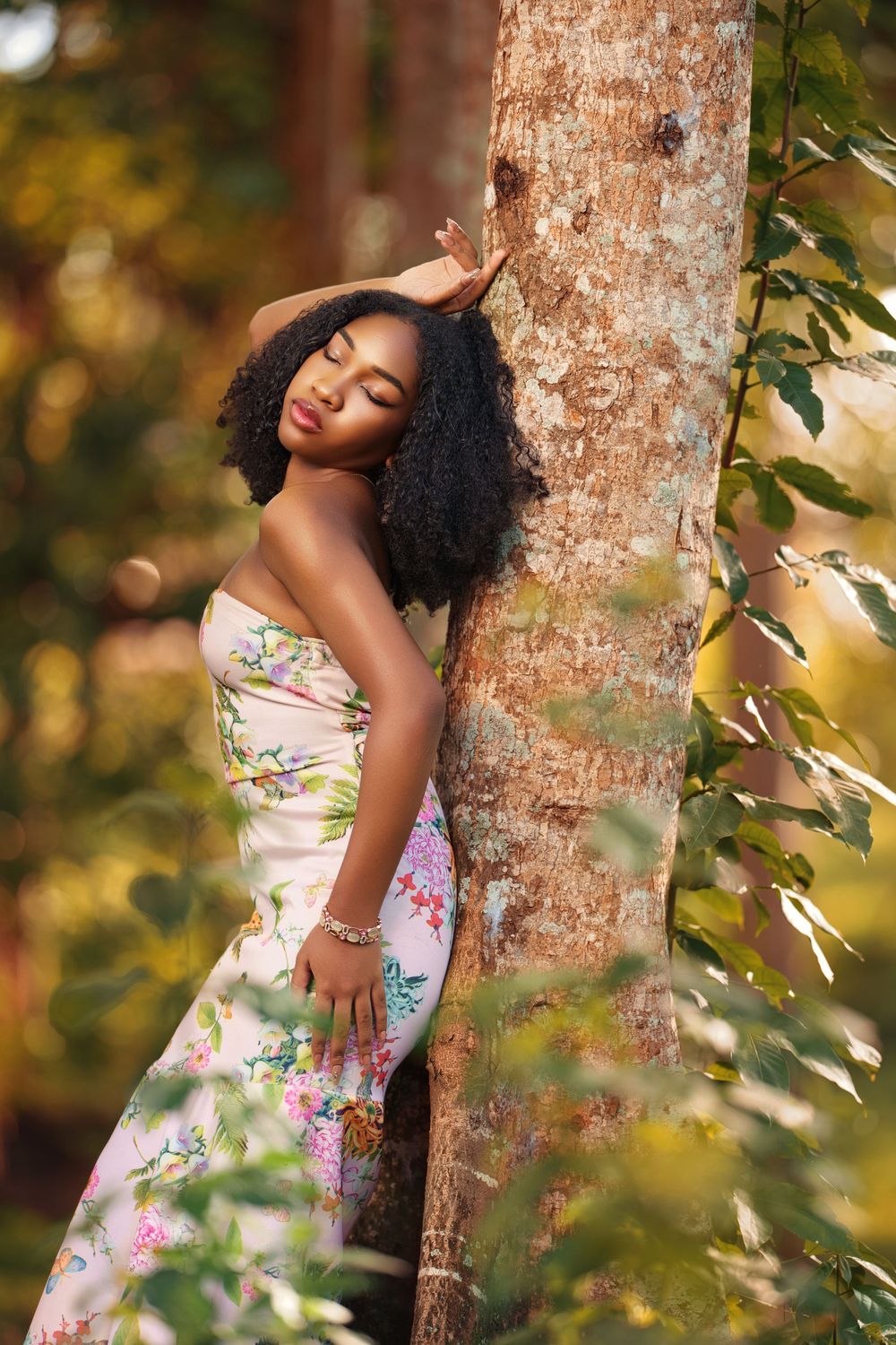 A woman in a floral dress poses gracefully against a tree trunk in a dreamy outdoor setting with golden sunlight.