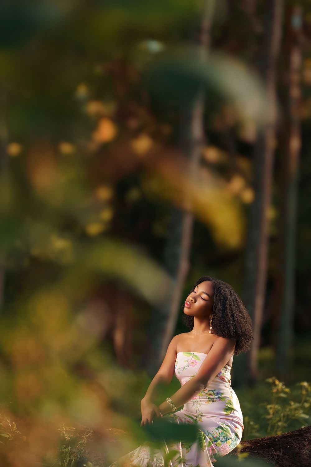 A woman in a white floral dress poses thoughtfully in a dreamy forest setting with soft bokeh effects in the background.