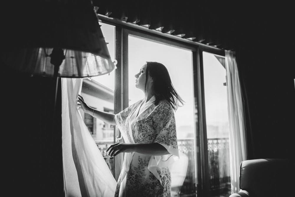 Novia mirando por la ventana durante los preparativos de la boda, luz suave y ambiente íntimo.