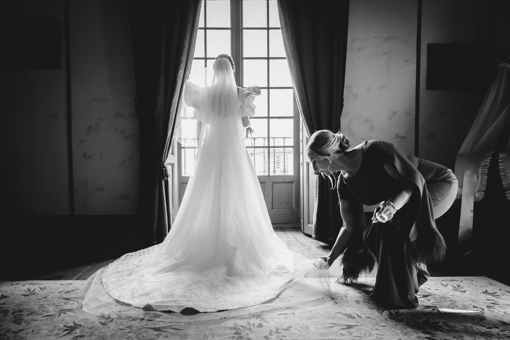 Novia de espaldas frente a la ventana con el vestido de boda colocado, preparativos en habitación elegante.