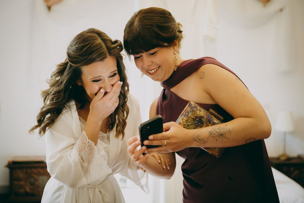Novia y amiga riendo mientras miran el móvil en los preparativos de la boda, momento espontáneo.