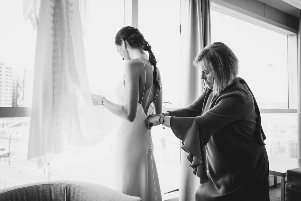 Madre ayudando a la novia a vestirse junto a la ventana, escena íntima de preparativos de boda.