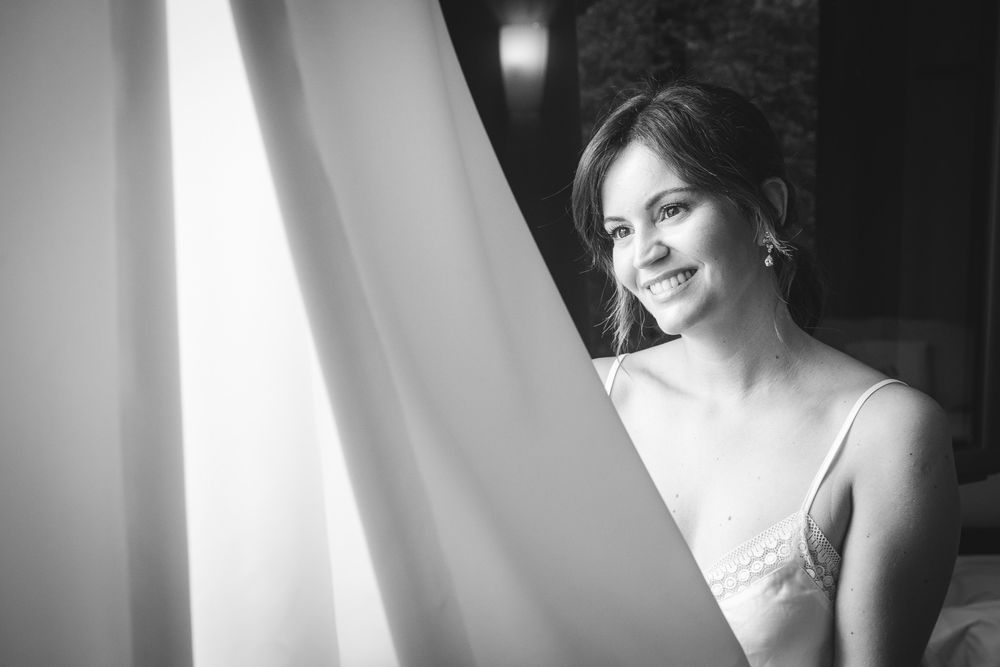 Novia sonriente junto a la ventana con el velo, fotografía natural de preparativos de boda.