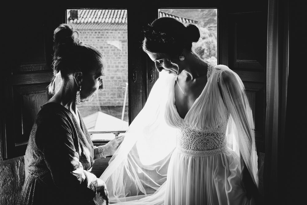 Novia ayudada por una familiar mientras se coloca el vestido junto a la ventana