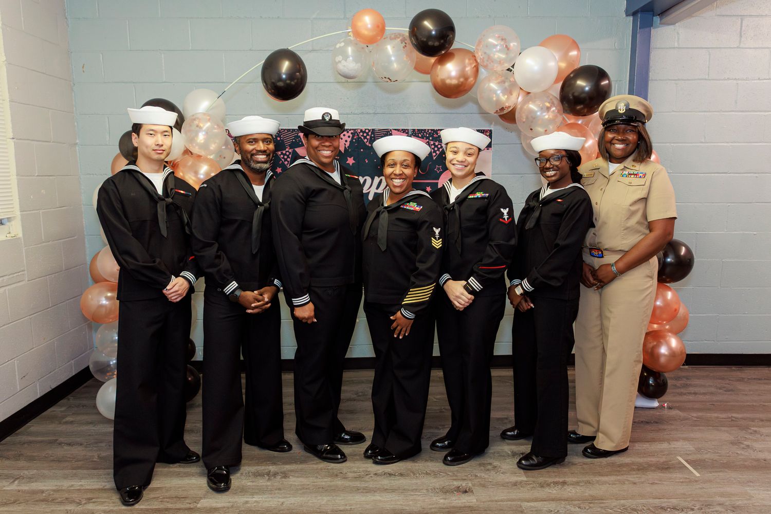 A group of people in formal black attire dance in a line at a celebration event with pink and black balloons.