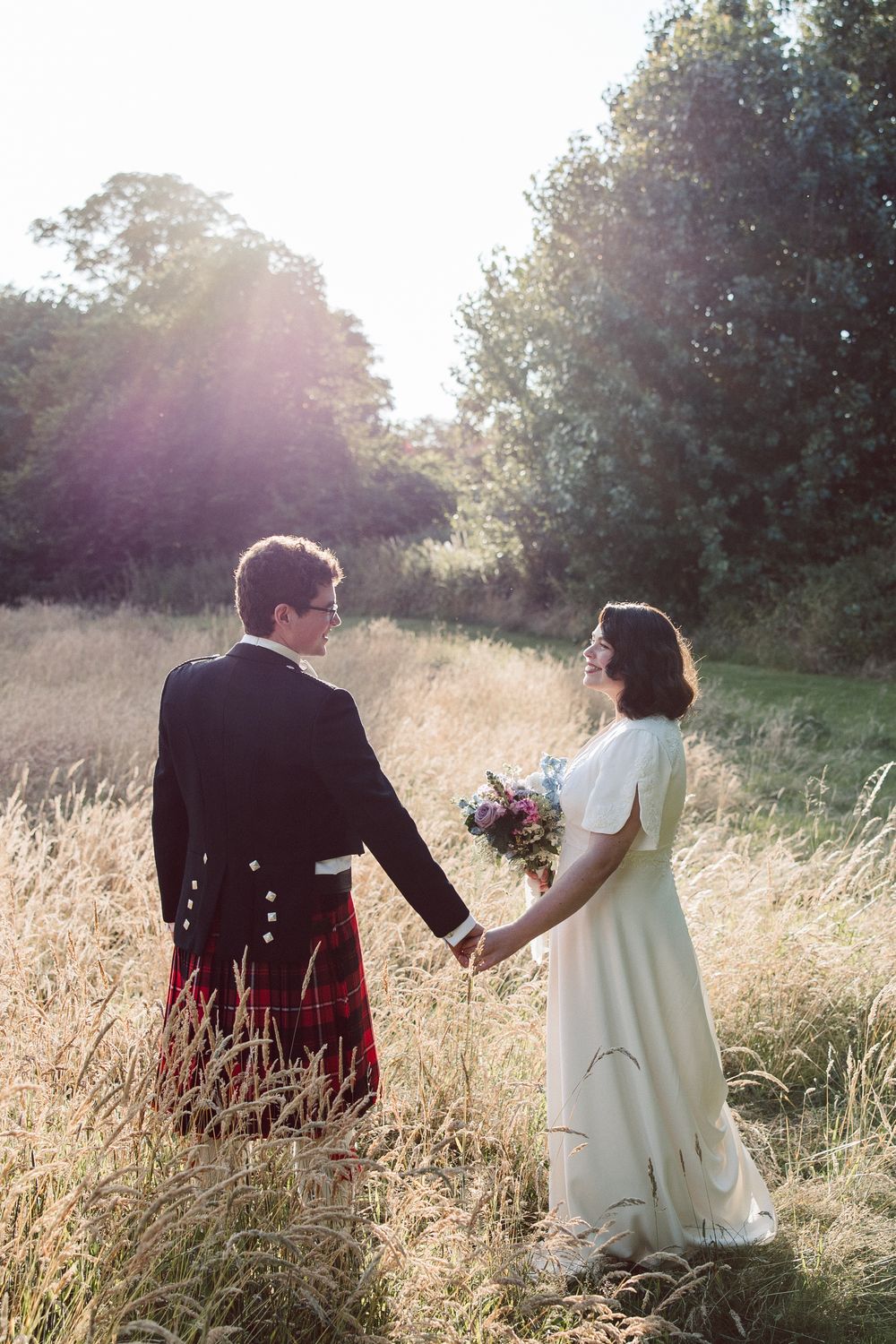 A couple sits together in a sunlit meadow of tall golden grass during sunset.