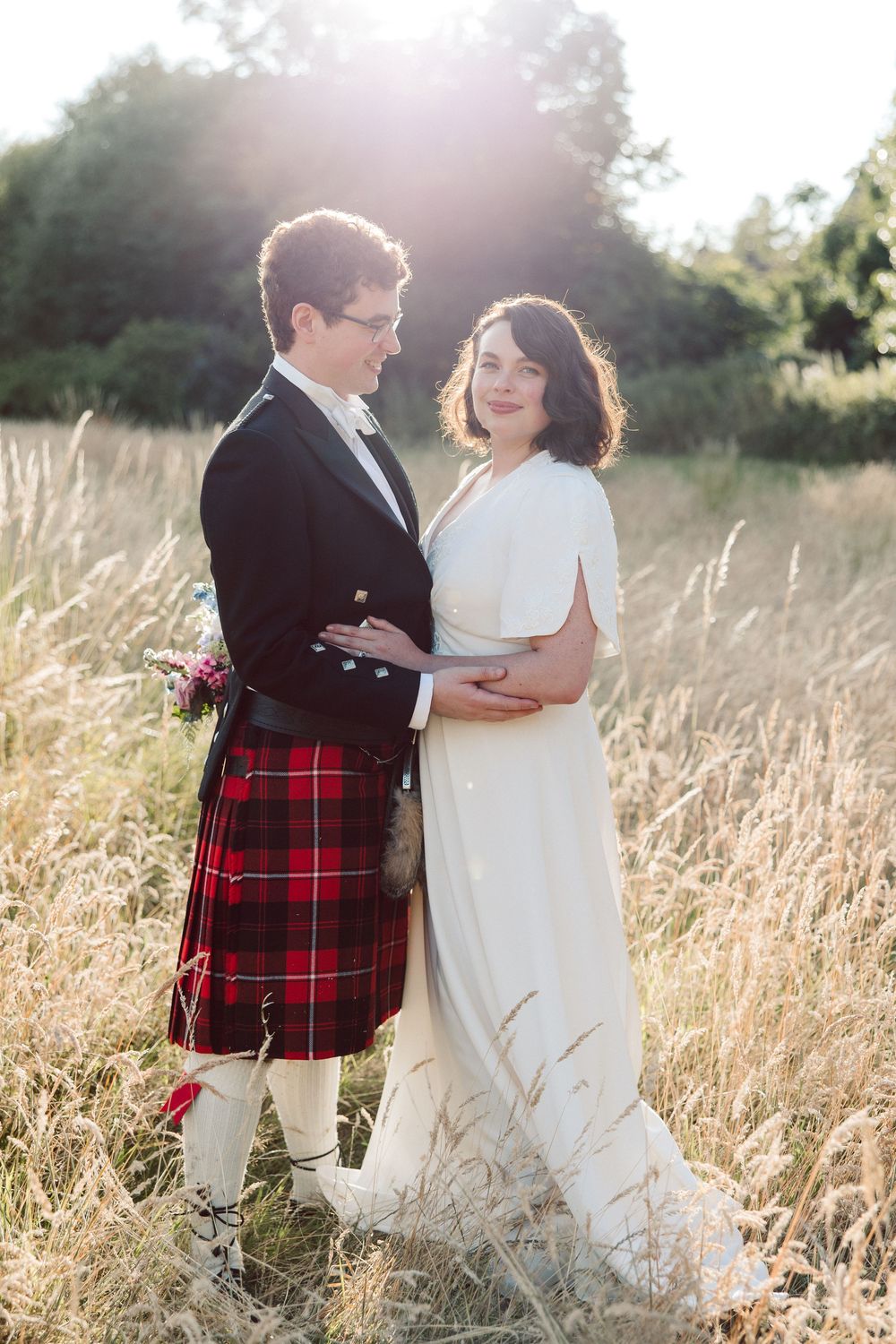 A couple poses in a sunlit meadow, with the groom wearing a traditional red tartan kilt and the bride in a flowing white dress.