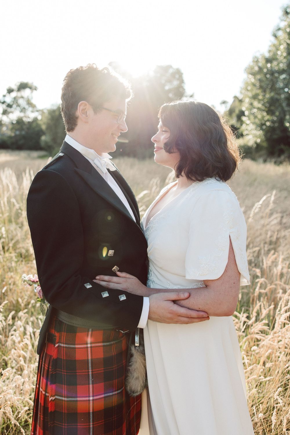 A romantic wedding couple embraces in a sunlit field wearing traditional Scottish attire with a red tartan kilt.