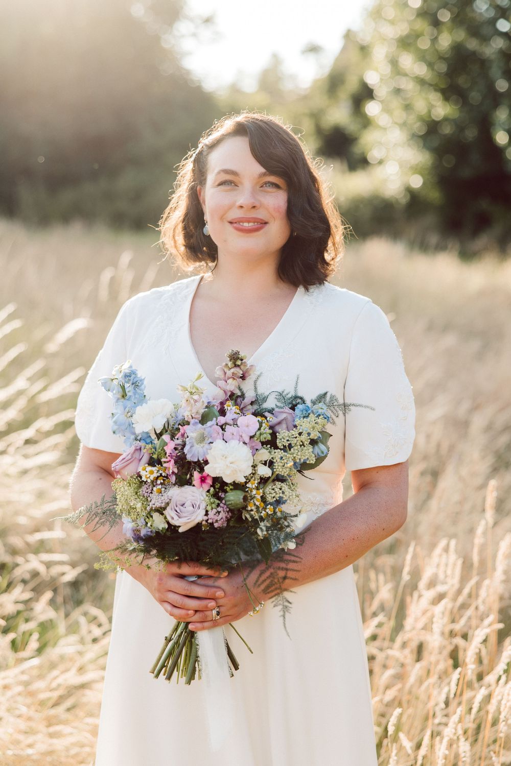 A person in a white dress holds a vibrant wildflower bouquet with purple and blue blooms in a sunlit field.