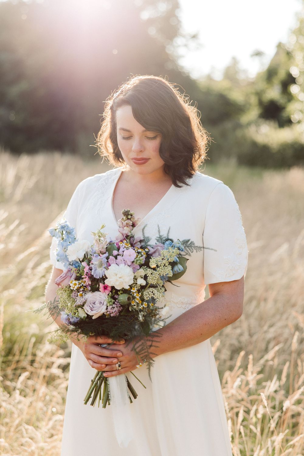 A person in a white dress holds a beautiful bouquet of pastel flowers in a sunlit field at golden hour.