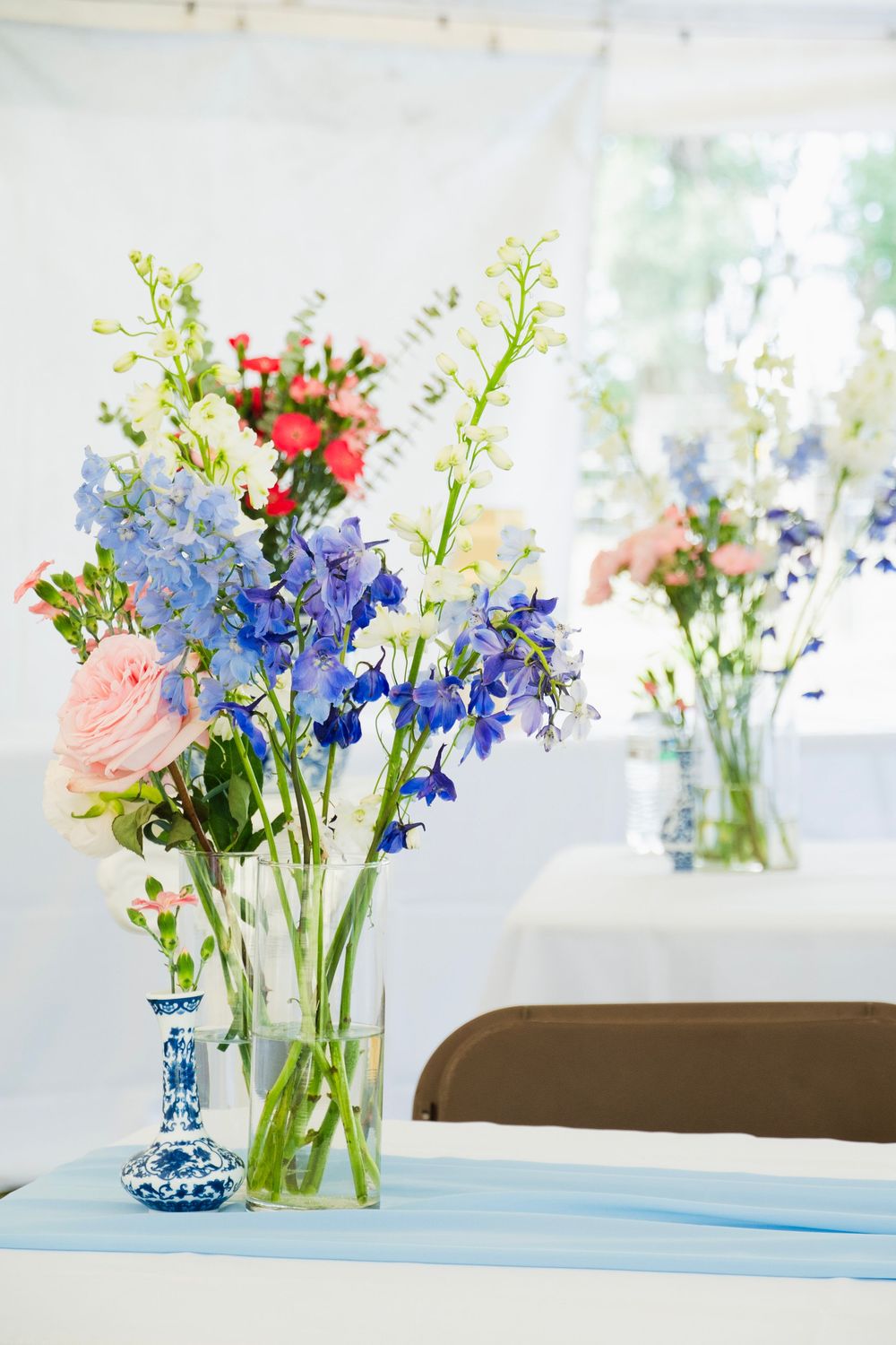 Tall glass vases with blue delphiniums and pink roses decorate tables inside a bright wedding tent.