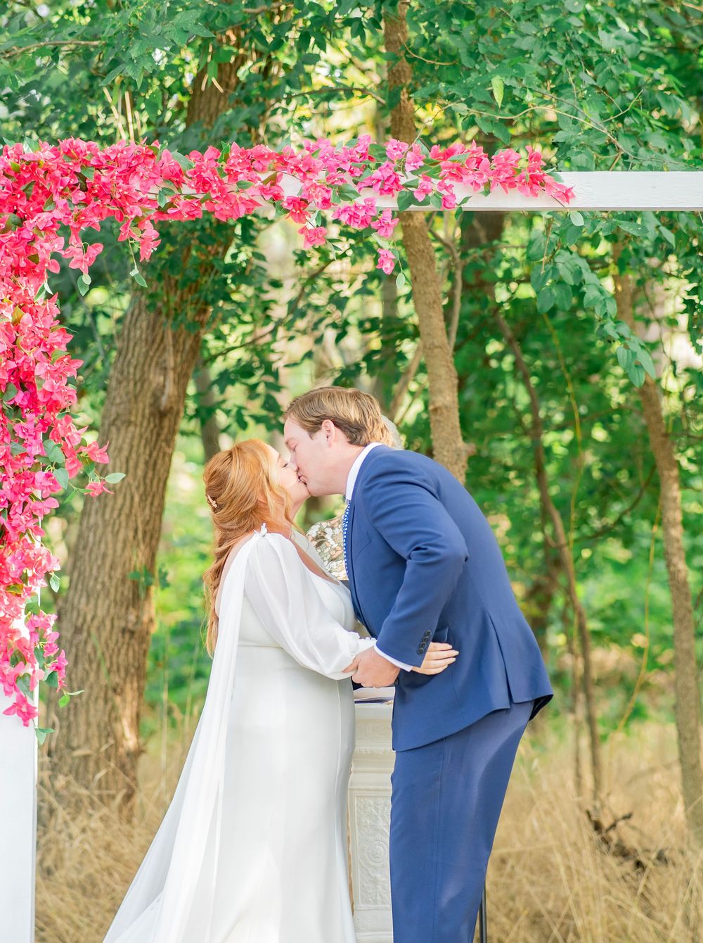Bride and groom share first kiss under pink bougainvillea arch at outdoor wedding ceremony.