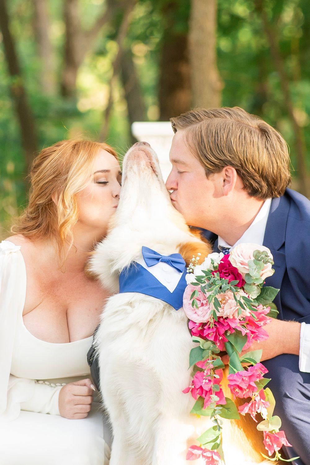 Bride and groom both kiss their dog wearing a blue bow tie bandana at outdoor wedding.