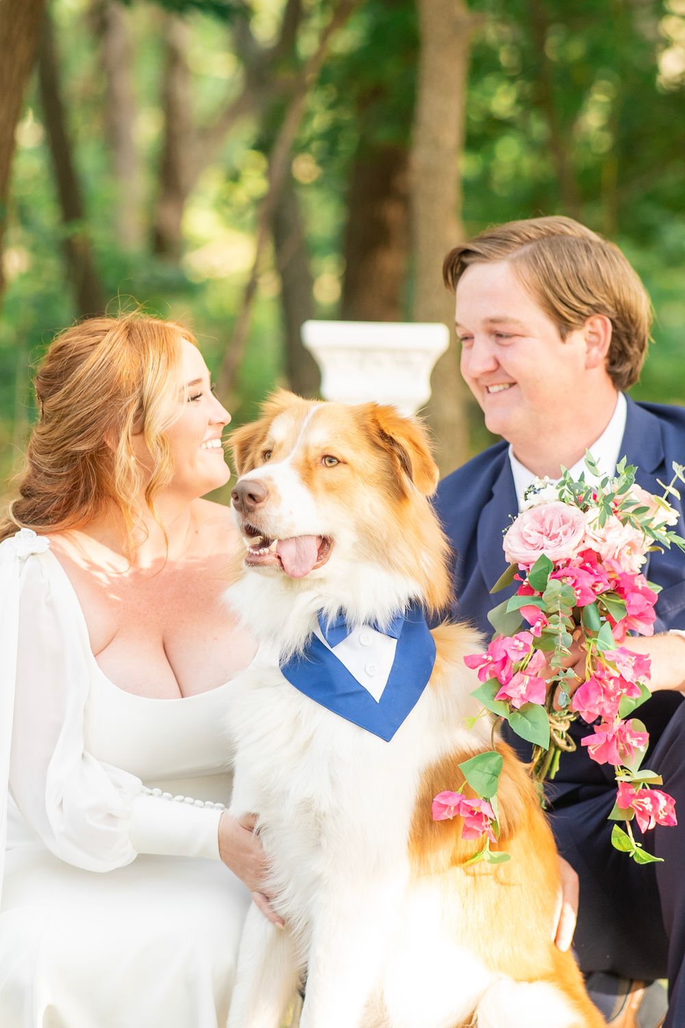 Smiling bride and groom pose with their happy Australian Shepherd dog wearing a blue bandana outdoors.