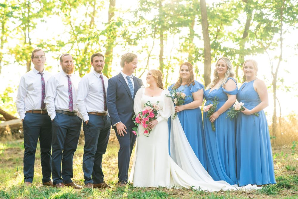 Wedding party poses in sunlit forest with bridesmaids in blue dresses and groomsmen in navy suits.