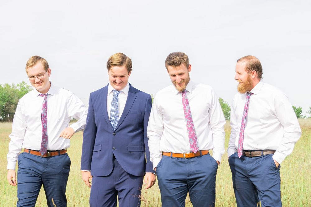 Four groomsmen in navy blue pants and pink ties walking together through a grassy field.