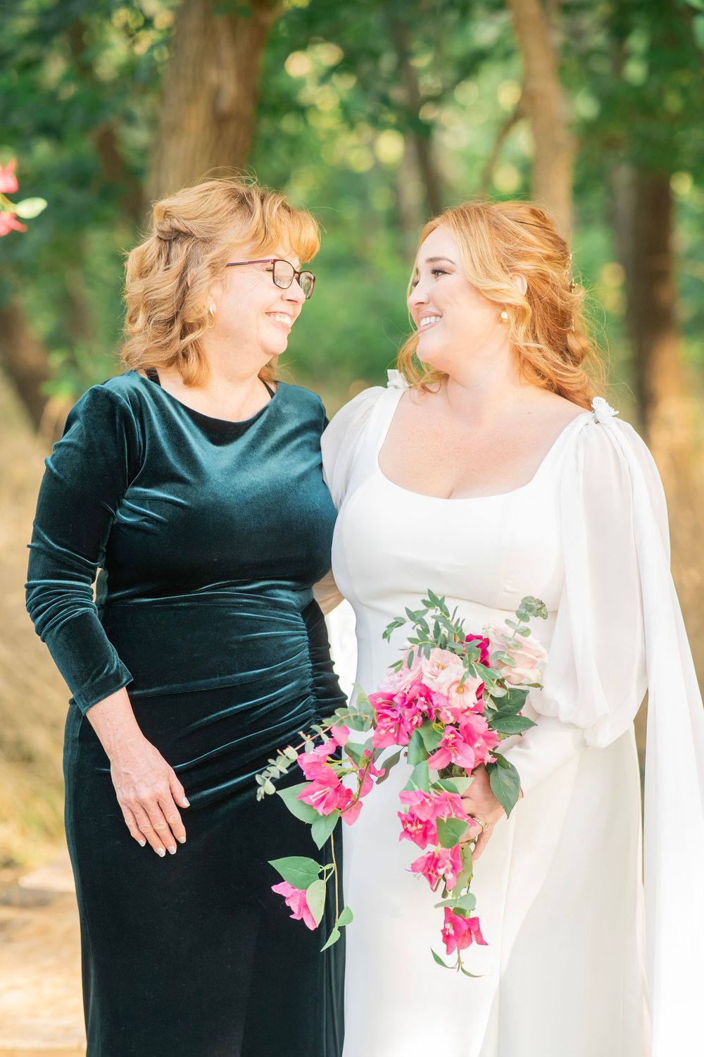 Bride in white gown with cascading pink bouquet shares a warm smile with her mother outdoors.