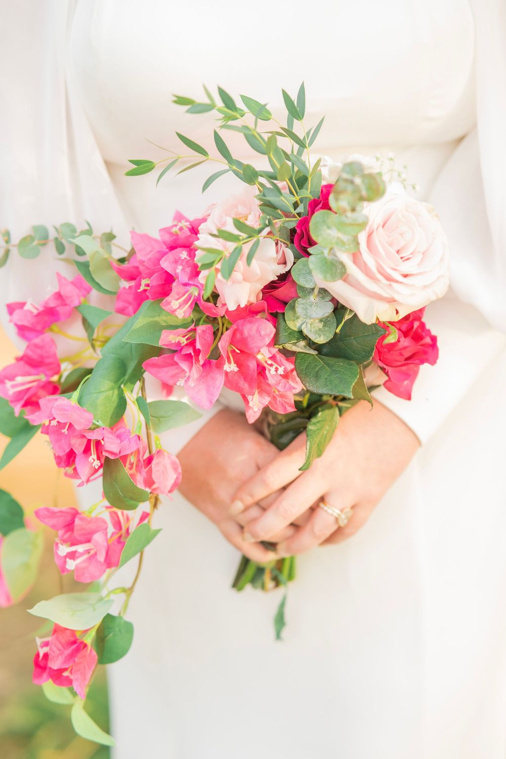 Close-up of bride holding cascading bouquet of hot pink bougainvillea and blush roses in white dress.