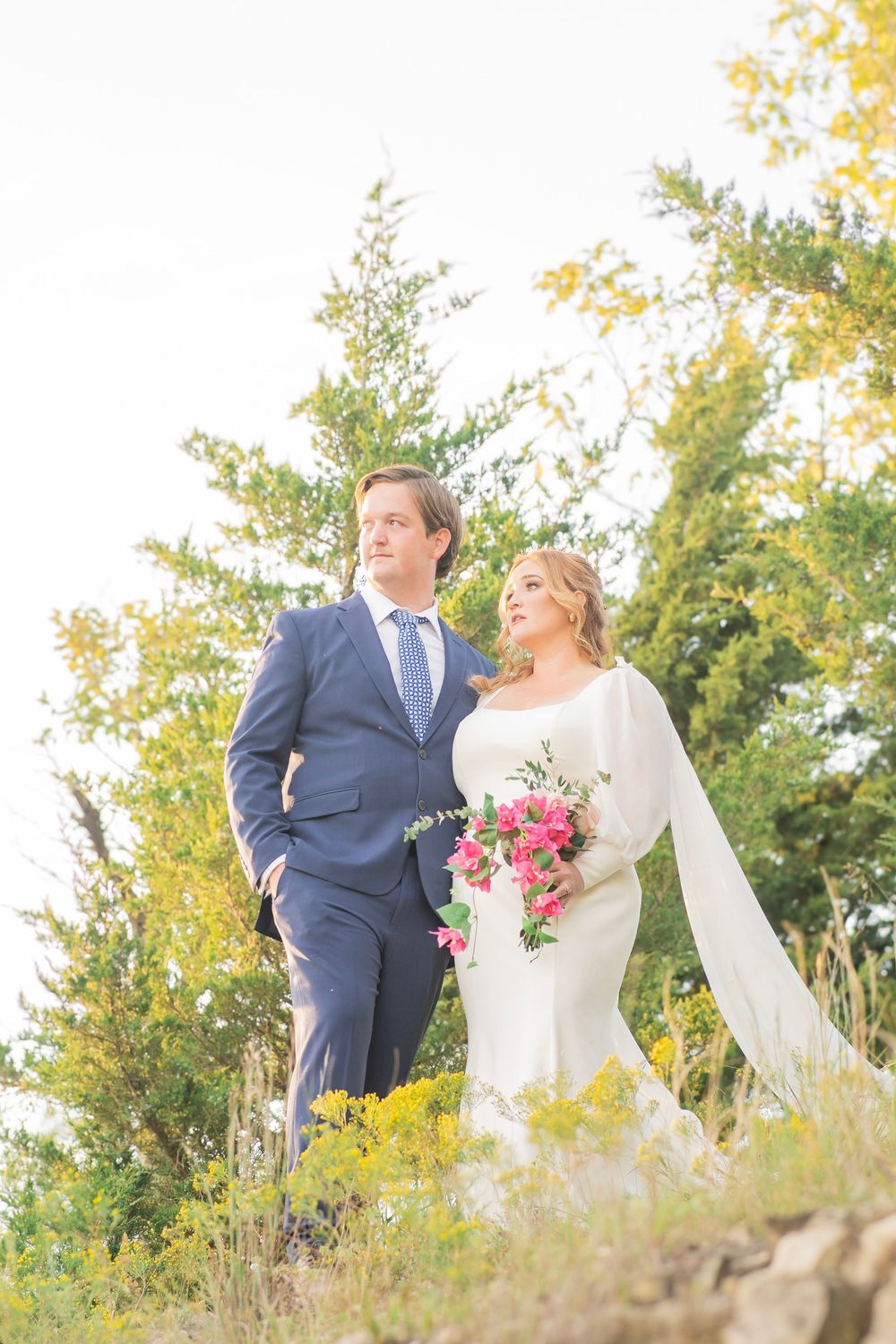 Wedding couple standing together in golden outdoor setting, bride in white gown holding pink bouquet, groom in navy suit.