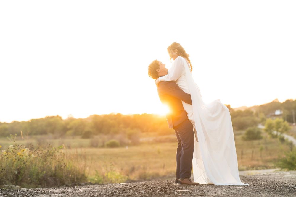 Groom lifting bride at sunset in an open field, bride in elegant white gown, romantic golden hour lighting.