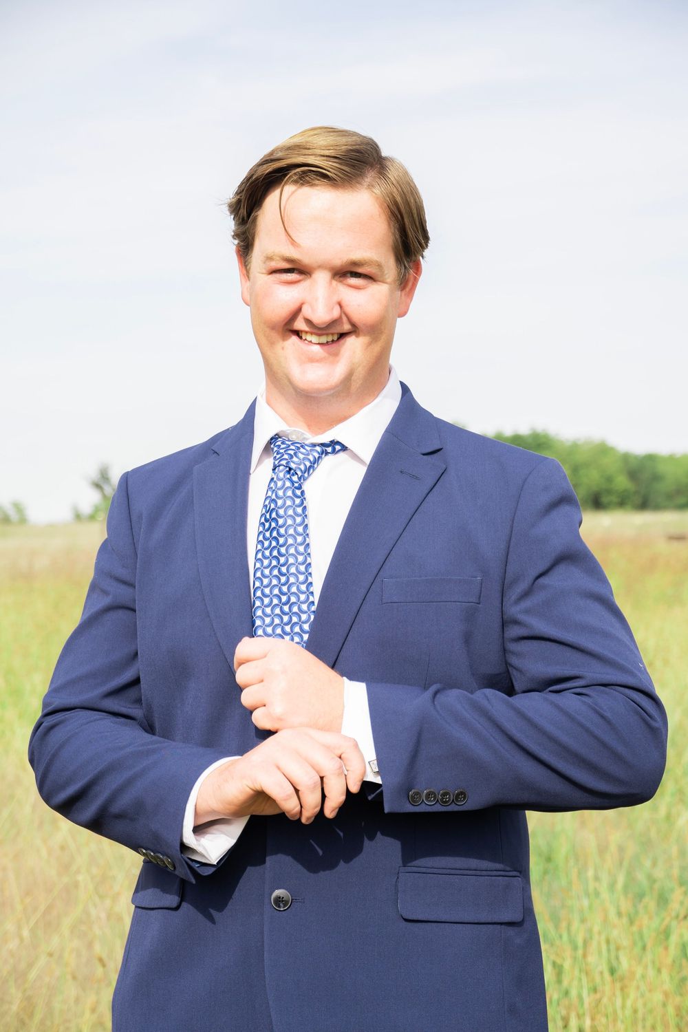 Happy groom in a navy suit adjusting his cufflinks while smiling outdoors in a field.