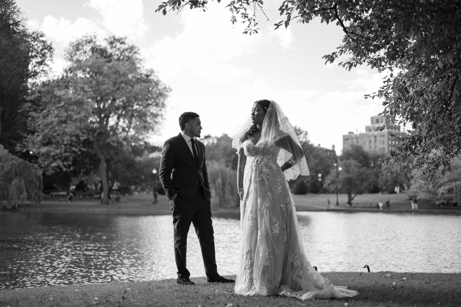A couple in wedding attire stands together by a tranquil pond in a scenic park setting, captured in black and white.