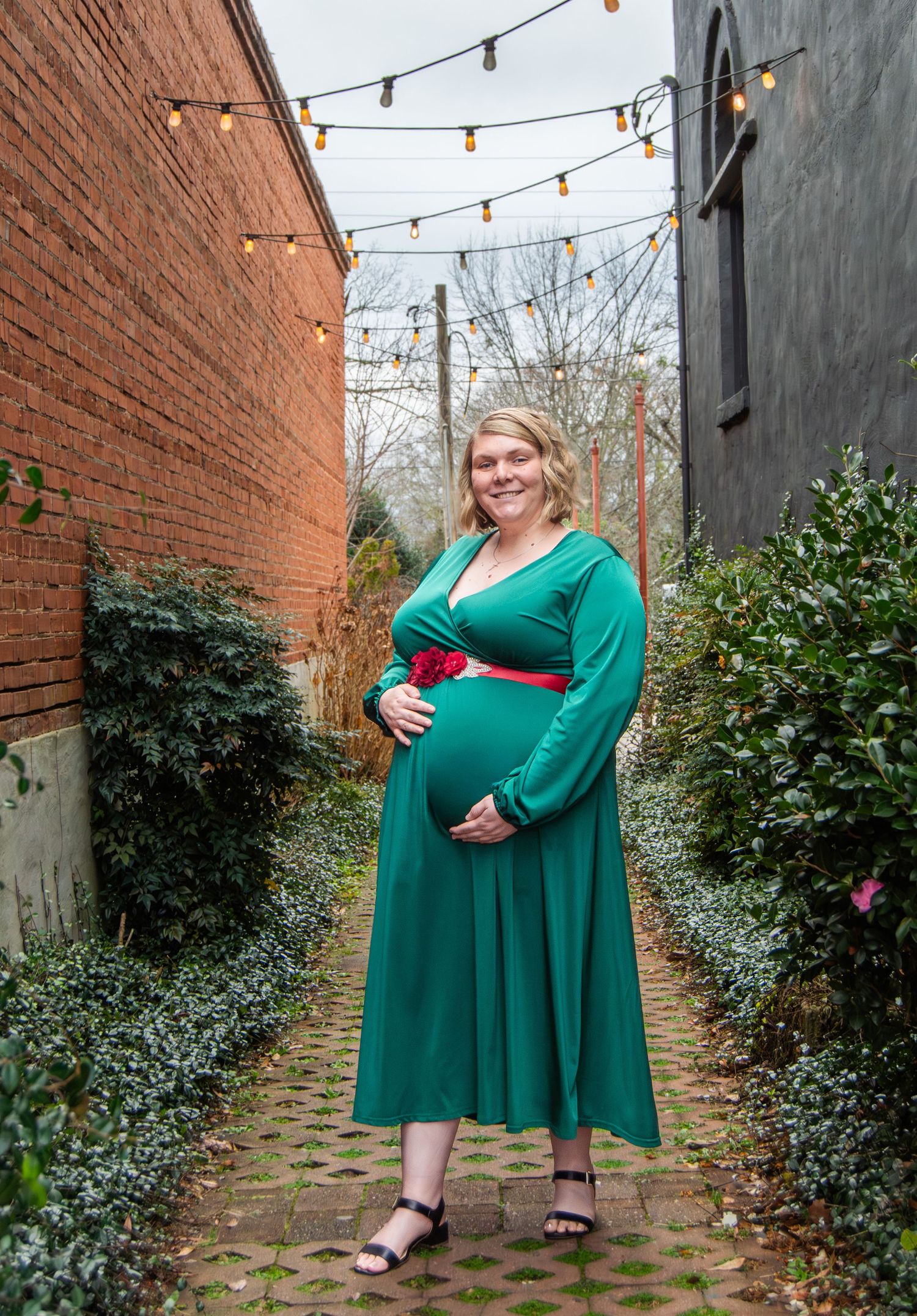 A person in a flowy emerald green dress poses in a brick alleyway decorated with string lights and greenery.
