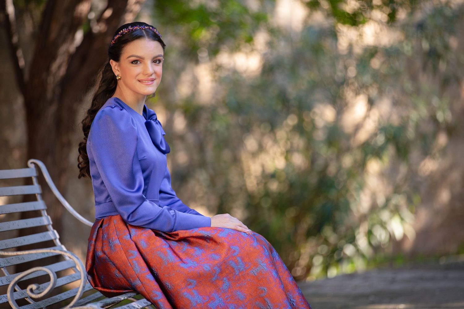 Woman in vintage blue and red dress sitting on garden bench.