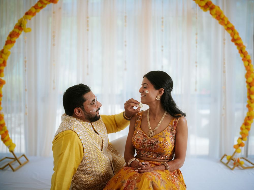 A couple shares a joyful moment during a pre-wedding celebration with orange marigold decor and white backdrop.