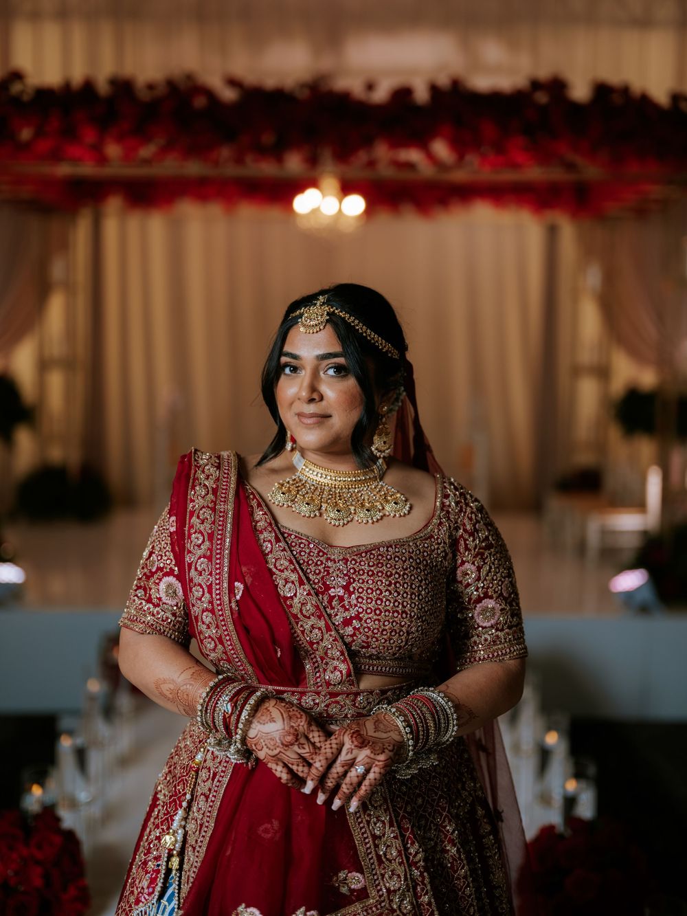 A bride in a traditional red and gold embroidered lehenga poses elegantly at an Indian wedding reception venue.