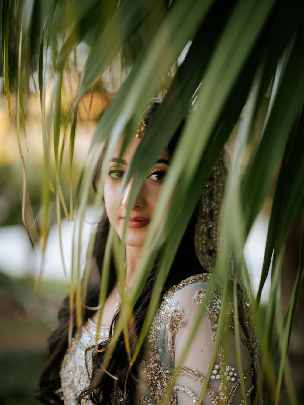 Artistic portrait through palm fronds showing a glittering formal dress in soft natural lighting.