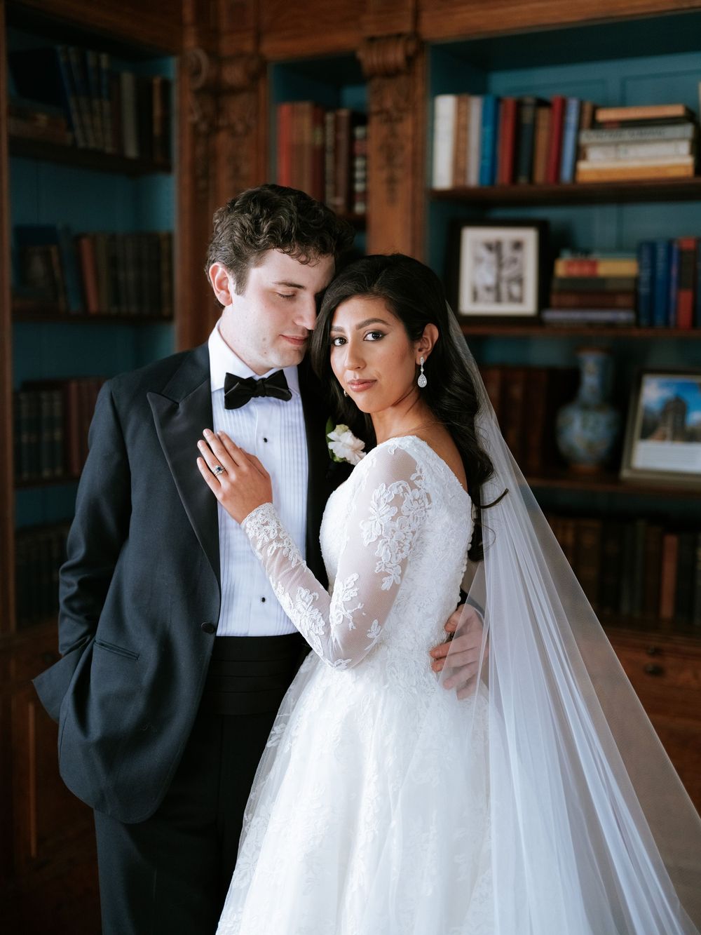 A romantic wedding couple embraces in a cozy library setting with bookshelves and warm lighting.