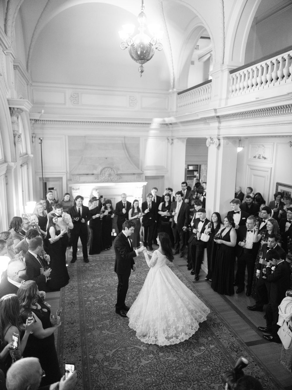 Elegant wedding reception dance in a grand ballroom with guests watching from the sides as couple twirls in white gown.