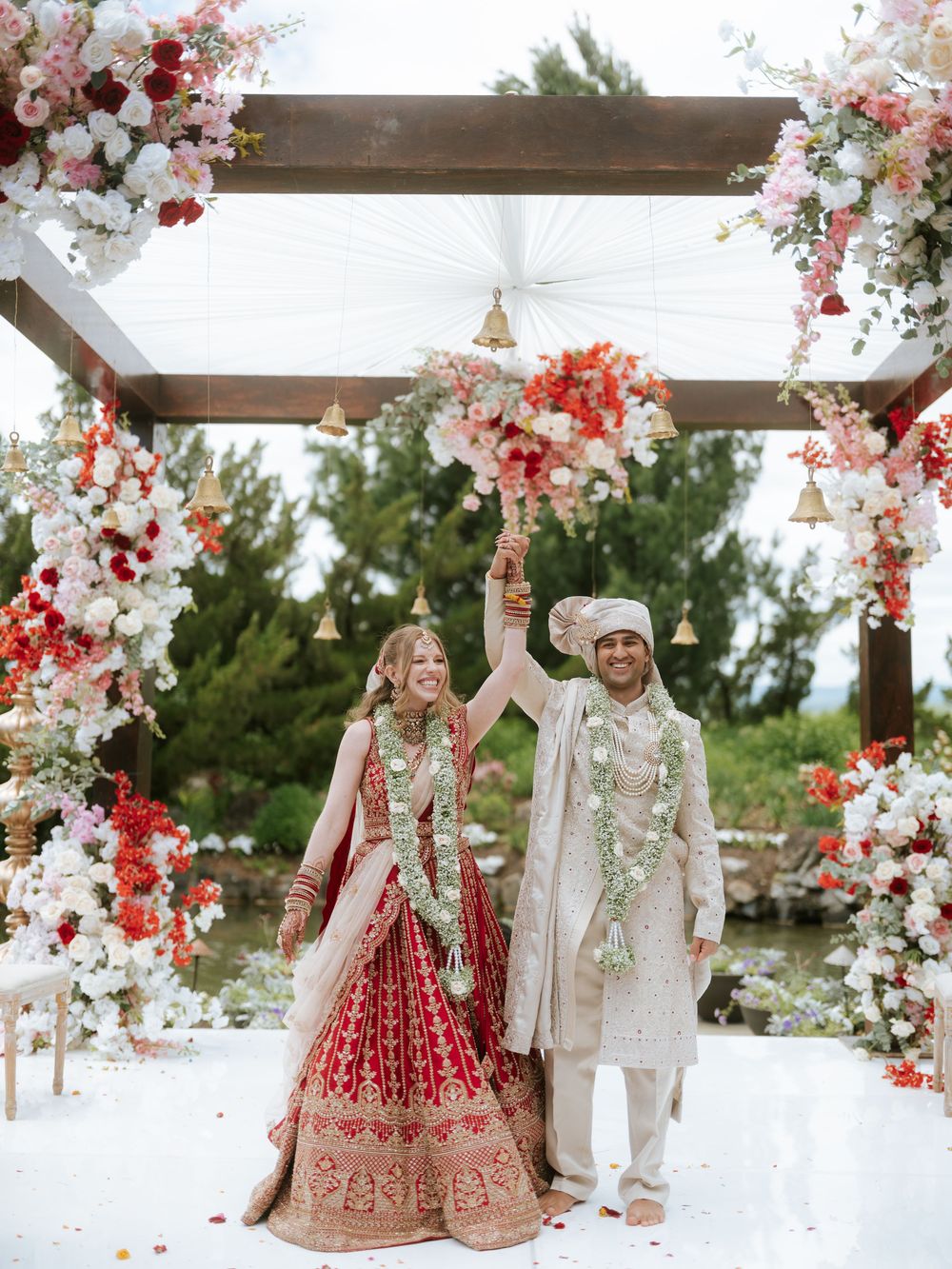 A joyful couple celebrates under a floral mandap, bride in red lehenga and groom in ivory sherwani, raising hands together.