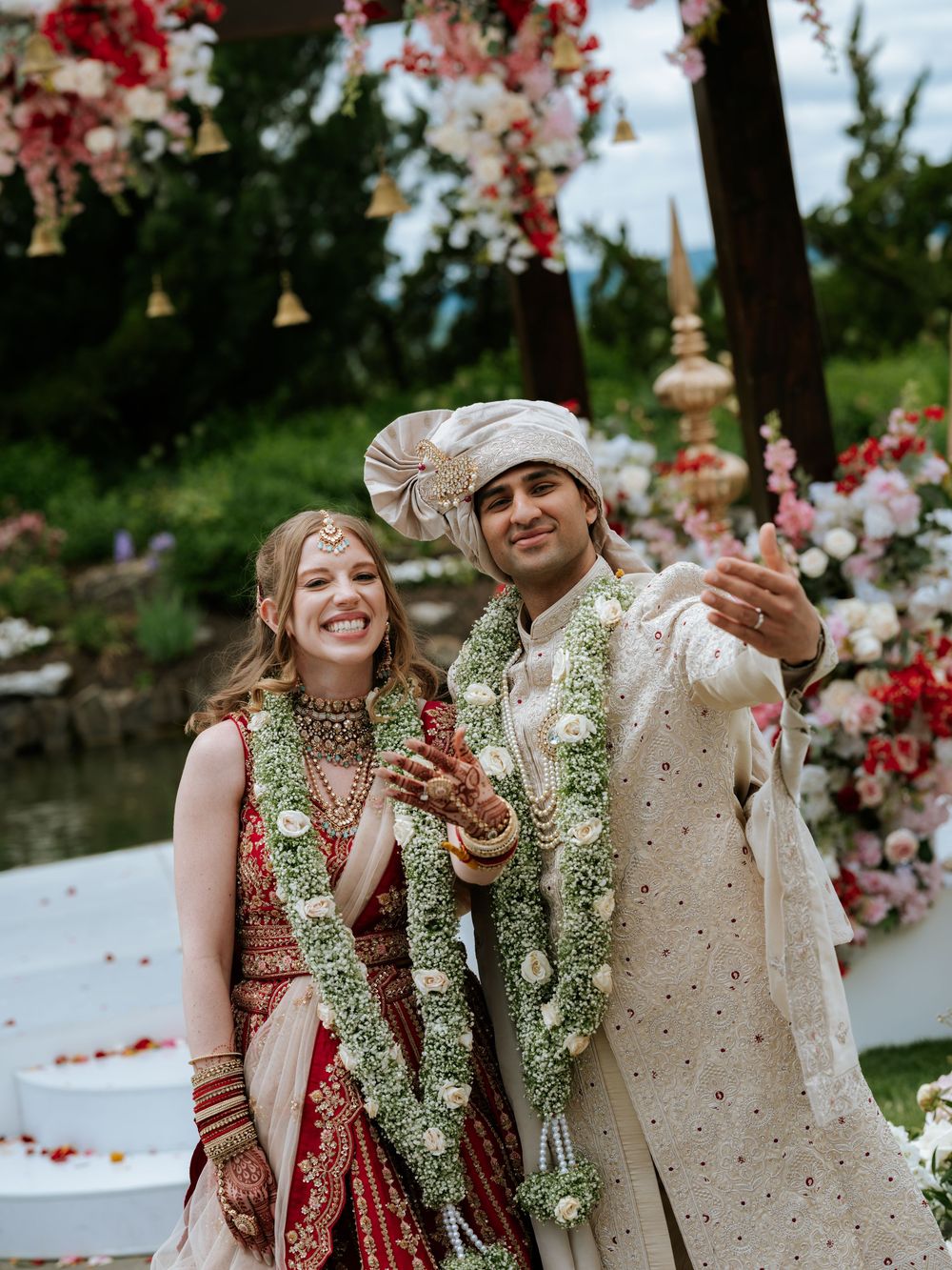 Smiling couple in traditional Indian wedding attire with floral garlands posing outdoors at decorated ceremony.