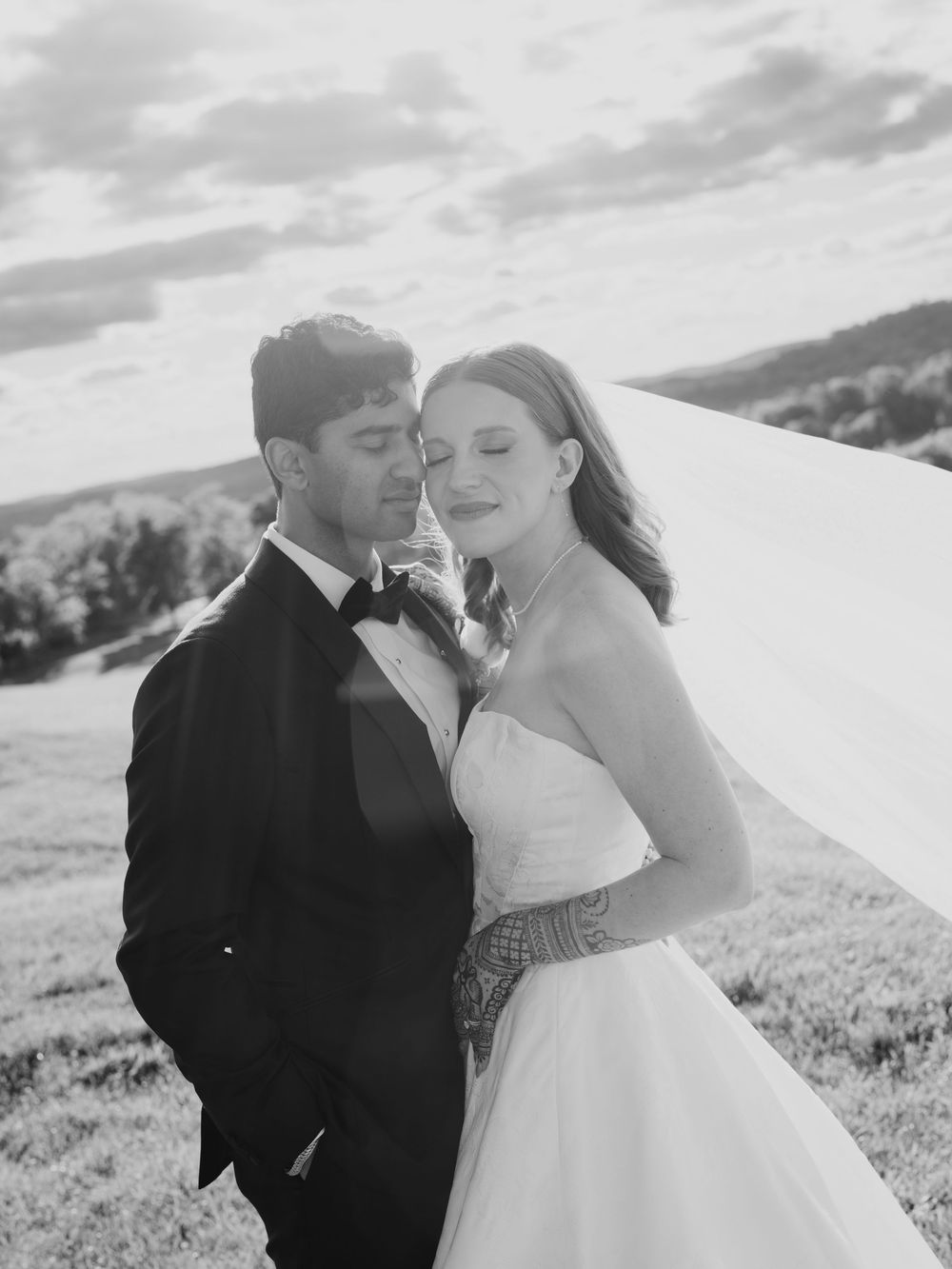 Black and white wedding photo of bride in strapless gown with veil and groom in tuxedo embracing outdoors.