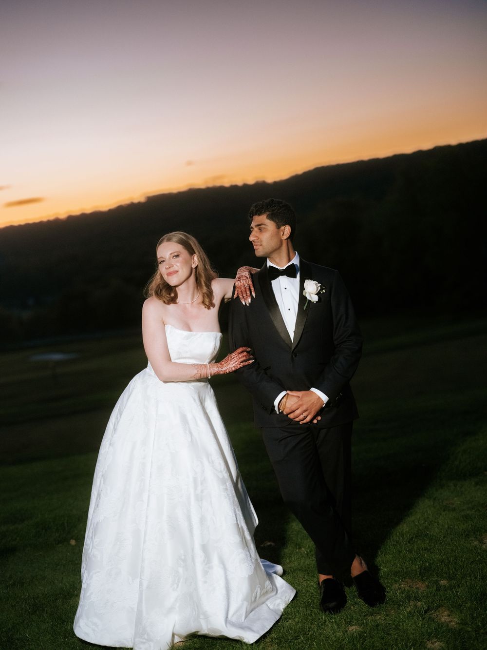 Bride in white strapless gown and groom in black tuxedo posing at sunset with scenic hillside view.