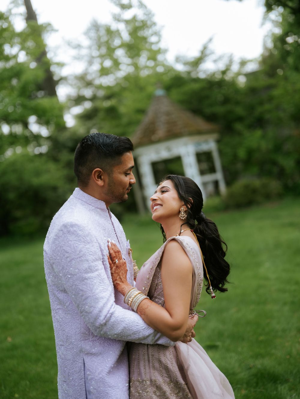 Indian couple in traditional wedding attire embracing and smiling in a lush garden with gazebo backdrop.