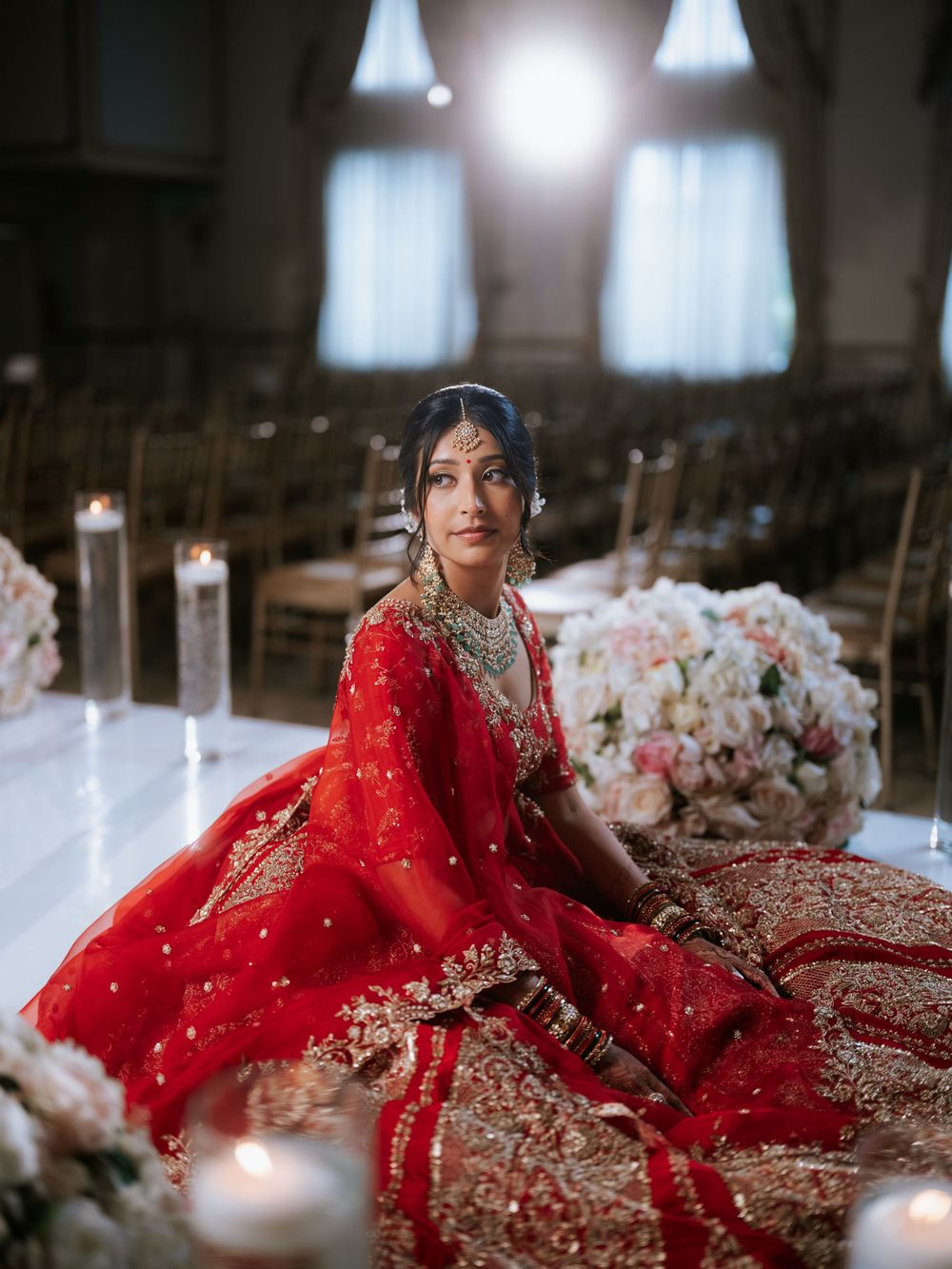 A bride in a stunning red and gold embroidered lehenga sits gracefully surrounded by candles and floral arrangements.