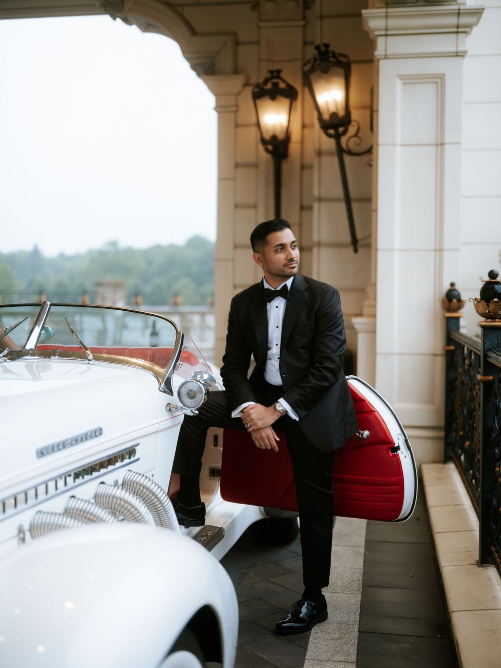 Elegant groom in black tuxedo leaning against a vintage white convertible car at a luxury estate venue.