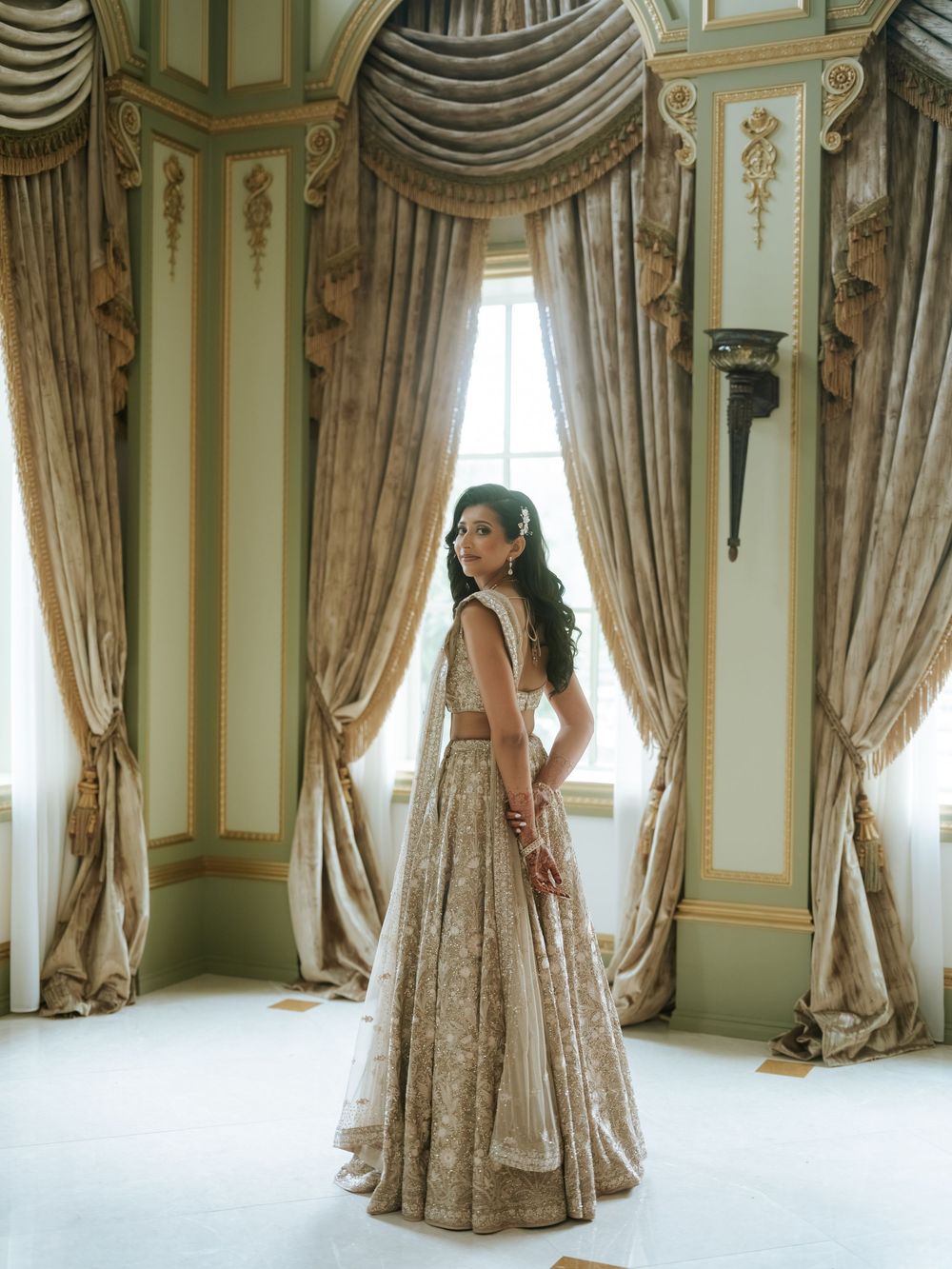 Bride in gold embroidered lehenga standing by ornate draped curtains in an elegant ballroom.