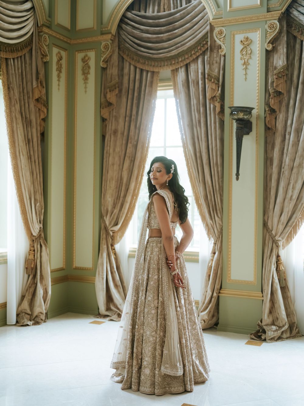 Bride in gold embroidered lehenga standing by ornate draped curtains in an elegant palace room.