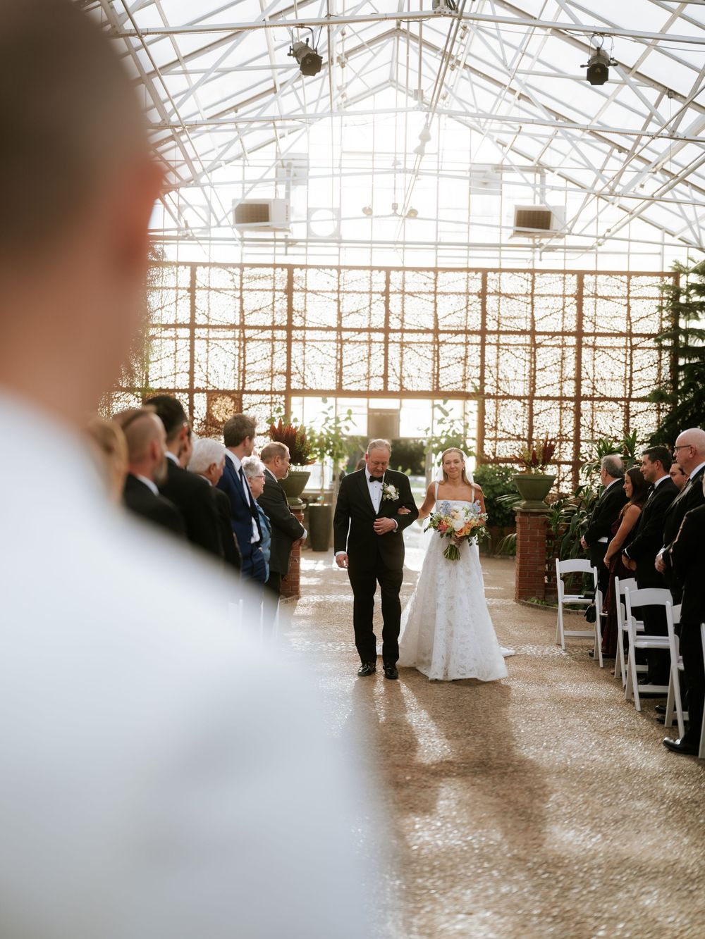 Bride walks down the aisle with escort in a sunlit greenhouse wedding venue filled with seated guests.