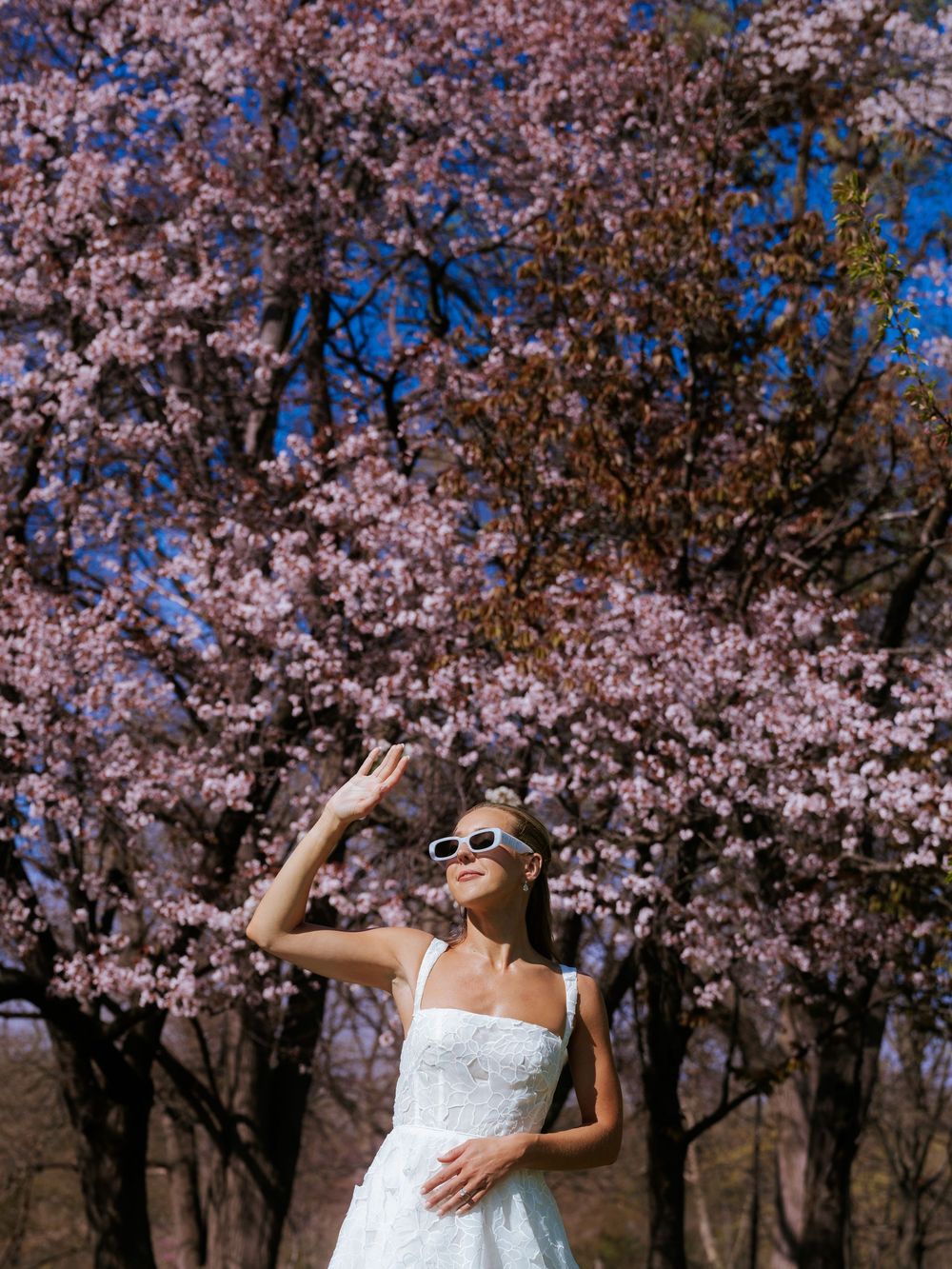 Woman in white dress and sunglasses posing before blooming pink cherry blossom trees under a blue sky.