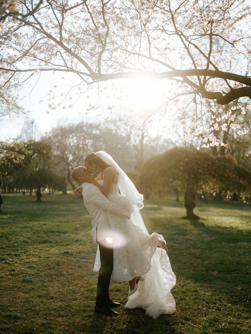 Bride and groom share a romantic kiss at golden hour in a sunlit park, with the bride's veil flowing gracefully.